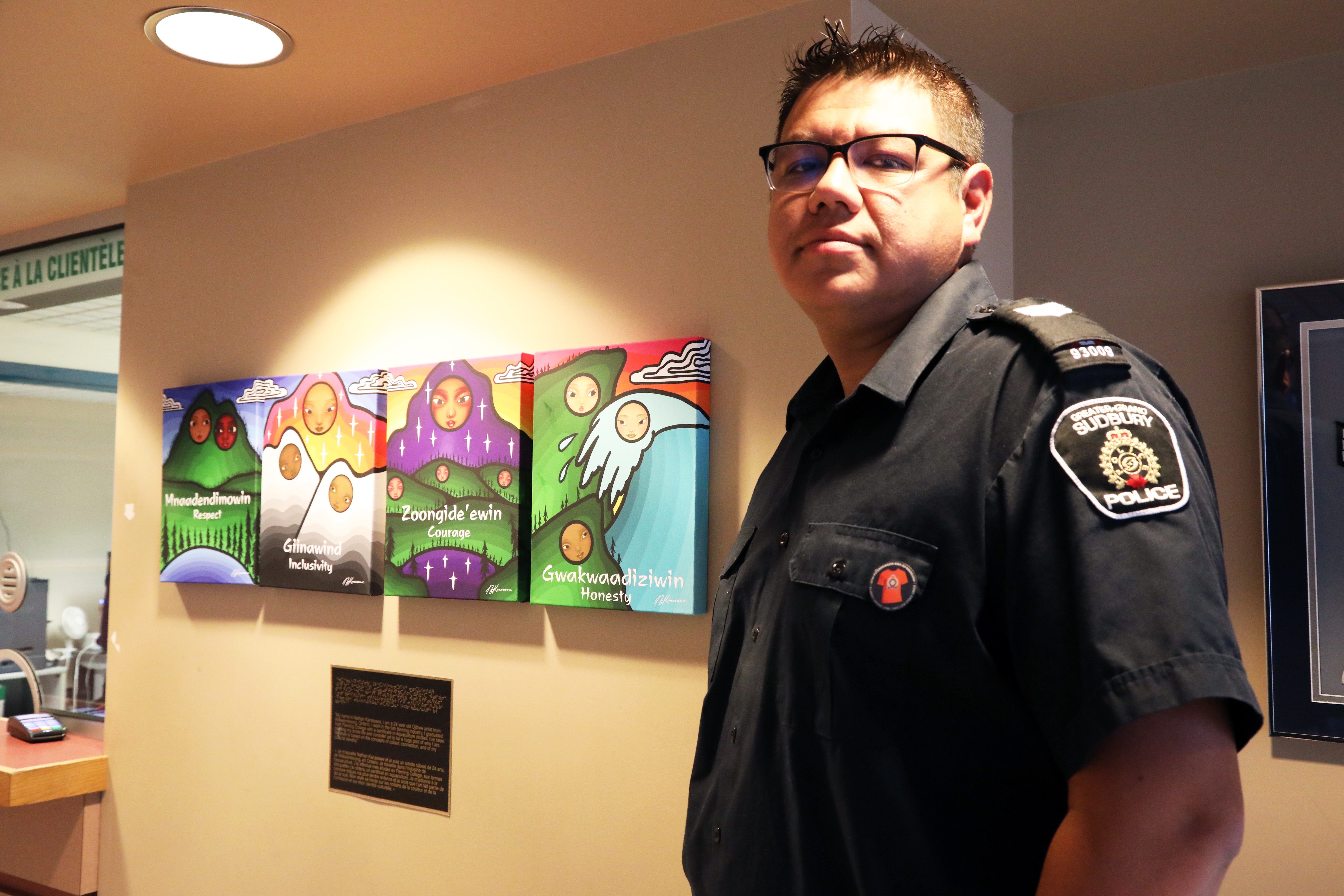 Sergeant Josh Rickard Officer wearing orange pin standing in front of Indigenous art work